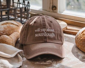 May include: A brown baseball cap with the embroidered text "will yap about sourdough" in white. The cap is on a wooden surface with loaves of bread and a white cloth. The cap has a curved brim and a classic six-panel design.