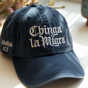 May include: Navy blue baseball cap with the embroidered text "Chinga la Migra" in white gothic lettering. The phrase "Abolish ICE" is embroidered on the side. The cap has a curved brim and a fabric strap closure.