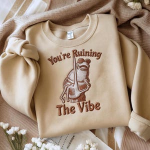 May include: Beige sweatshirt with the embroidered phrase "You're Ruining The Vibe" and a frog wearing sunglasses. The frog is holding a long stick. The sweatshirt is folded on a beige blanket.