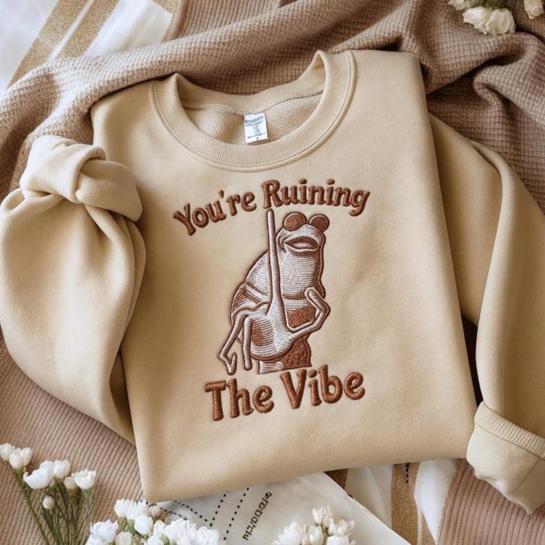 May include: Beige sweatshirt with the embroidered phrase "You're Ruining The Vibe" and a frog wearing sunglasses. The frog is holding a long stick. The sweatshirt is folded on a beige blanket.