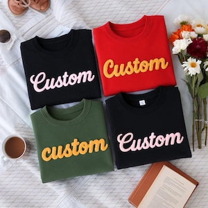 May include: Four crewneck sweatshirts in black, red, olive green, and black. Each sweatshirt has the word "Custom" embroidered across the front in a contrasting color. The sweatshirts are displayed on a white surface with flowers and accessories.