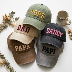 May include: A collection of five baseball caps in various colors including olive green, beige, brown, charcoal gray, and black. Each cap features embroidered text on the front panel reading POPS, DAD, PAPA, DADDY, and PAPI in contrasting thread colors.