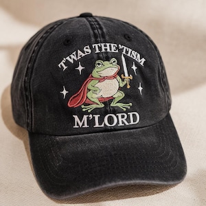 May include: Black baseball cap with embroidered design. The design features a green frog wearing a red cape and holding a sword, with the text "T'WAS THE 'TISM M'LORD" in white.