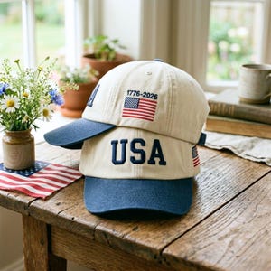 May include: Two beige baseball caps with navy blue brims are stacked on a wooden table. The top cap features the text "1776-2026" and a small American flag. The bottom cap displays the word "USA" in navy blue.