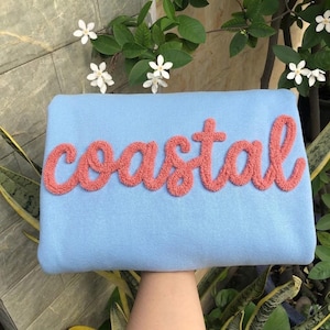 May include: A light blue sweatshirt featuring the word coastal in a textured, coral-colored, fuzzy, raised script font across the center. The garment is made of a smooth fabric and displays a distinct, tactile, embroidered-style lettering design.