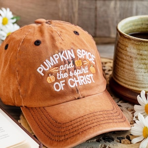 May include: An orange baseball cap with the text "PUMPKIN SPICE and the spirit OF CHRIST" in white. The cap has a curved brim and black accents. The image also shows a mug and flowers.