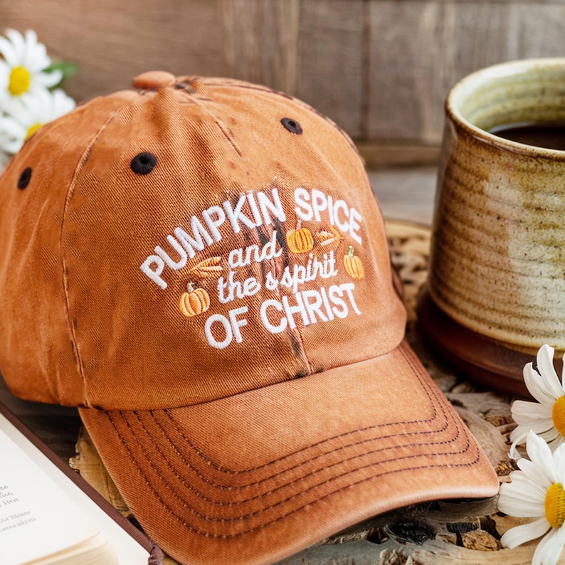 May include: An orange baseball cap with the text "PUMPKIN SPICE and the spirit OF CHRIST" in white. The cap has a curved brim and black accents. The image also shows a mug and flowers.