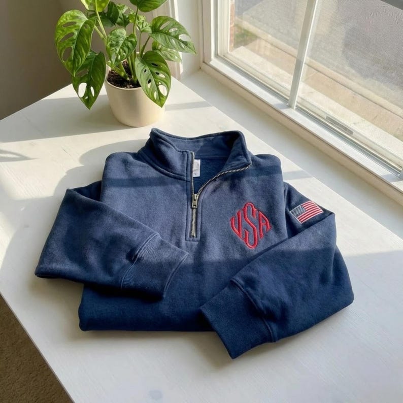 May include: Navy blue quarter-zip sweatshirt with a red "USA" monogram on the chest and a small American flag patch on the sleeve. The sweatshirt has a silver zipper and is folded neatly on a white surface.