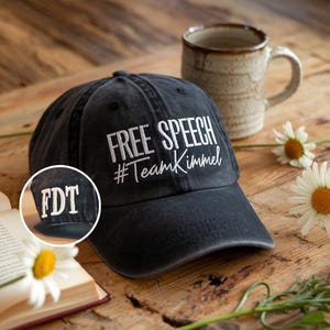 May include: A black baseball cap with the words "FREE SPEECH #TeamKimmel" embroidered in white. The cap also has the letters "FDT" embroidered on the side. A ceramic mug and daisies are in the background.