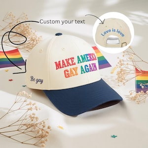 May include: A baseball cap with a cream-colored crown and a navy blue brim. The front of the cap features the text "MAKE AMERICA GAY AGAIN" in rainbow colors. The side says "Be gay" and the back says "Love is love".