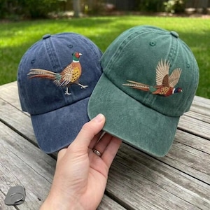 Puede incluir: Dos gorras de b&eacute;isbol, una azul marino y otra verde bosque, cada una con un bordado detallado de un fais&aacute;n. La gorra azul muestra un fais&aacute;n de pie, mientras que la verde presenta un fais&aacute;n en vuelo con plumaje de tonos marrones, dorados y rojos.