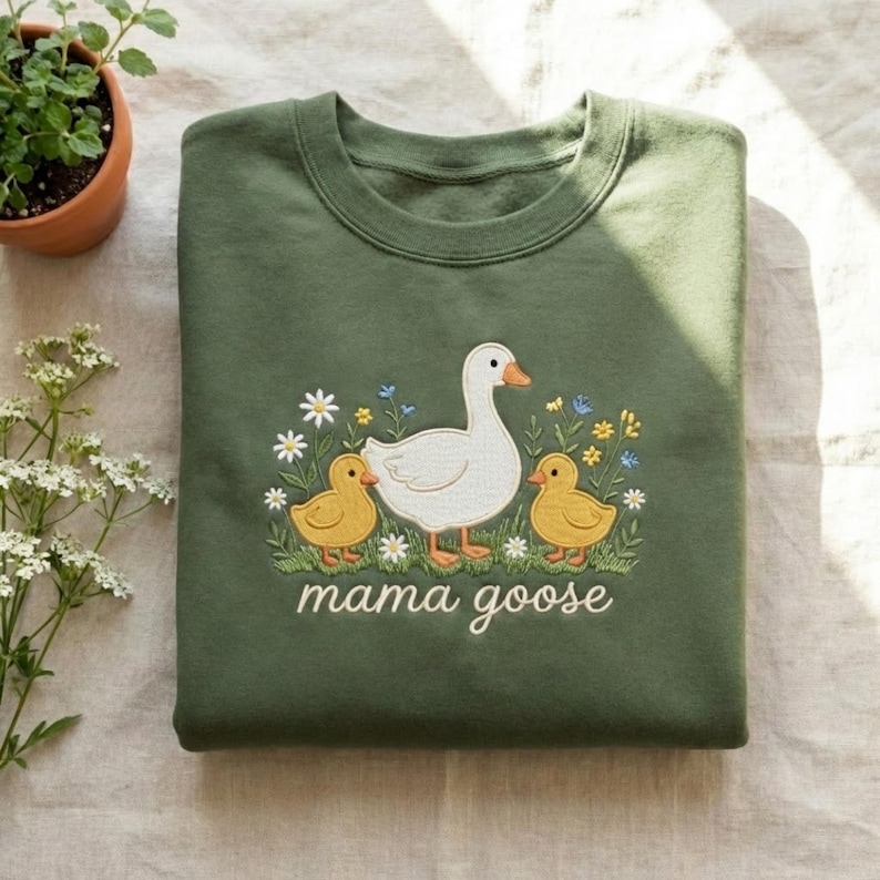 May include: An olive green sweatshirt featuring an embroidered design of a white goose, two yellow ducklings, and floral accents. The words "mama goose" are embroidered below the design. The sweatshirt is folded.