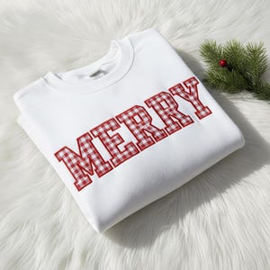 May include: A white sweatshirt with the word "MERRY" embroidered in red and white gingham letters. A sprig of evergreen with a red berry is in the upper right corner. The sweatshirt is folded on a white, fluffy surface.
