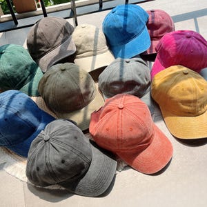 Puede incluir: Una colecci&oacute;n de doce gorras de b&eacute;isbol en varios colores, incluyendo gris oscuro, verde bosque, azul marino, beige, azul brillante, granate, magenta, gris, amarillo mostaza y coral. Cada gorra presenta un dise&ntilde;o de seis paneles.