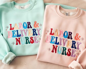May include: Two crewneck sweatshirts, one mint green and one light pink, embroidered with the words "LABOR & DELIVERY NURSE" in colorful, retro-style lettering. The sweatshirts have a soft, fleecy interior.