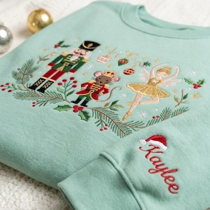 May include: A light green sweatshirt featuring an embroidered design of a nutcracker, a mouse in a red uniform, and a ballerina surrounded by pine branches and ornaments. The sleeve is embroidered with the name Kaylee and a small Santa hat.