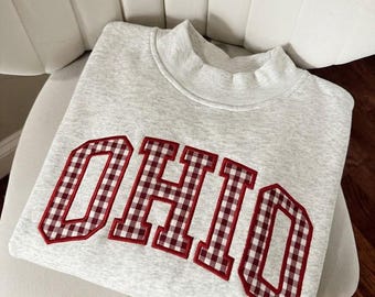 May include: A light gray heathered sweatshirt featuring the word OHIO in large block letters. The letters are filled with a red and white gingham pattern and are outlined with a thick red border. The garment has a mock neck collar.