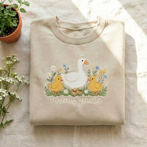 May include: Beige sweatshirt with an embroidered design featuring a white goose, two yellow ducklings, and floral accents. The phrase "mama goose" is embroidered below the design. The sweatshirt is folded neatly.