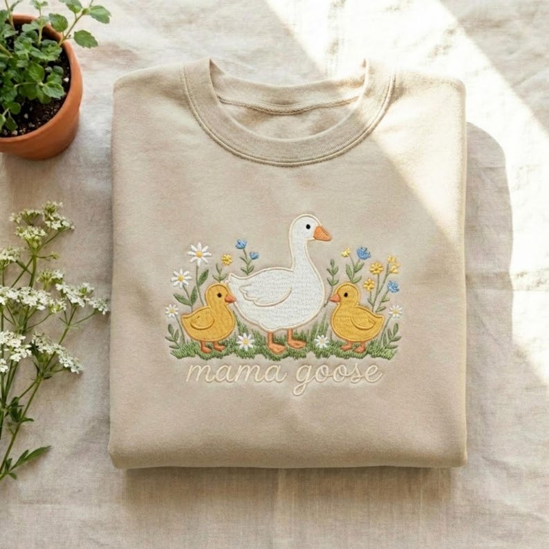 May include: Beige sweatshirt with an embroidered design featuring a white goose, two yellow ducklings, and floral accents. The phrase "mama goose" is embroidered below the design. The sweatshirt is folded neatly.