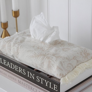 May include: A white tissue box cover with a beige floral pattern and white fringe sits on top of two books. The top book has the title "Leaders in Style" printed on the spine. The bottom book has the title "Abigail Ahern Masterclass" printed on the spine.