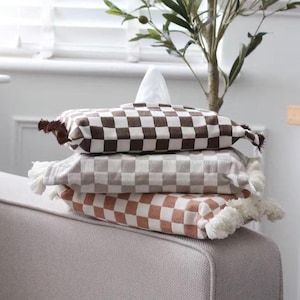 May include: A stack of three tissue box covers with a checkered pattern. The top cover is brown and white, the middle cover is grey and white, and the bottom cover is pink and white. Each cover has a fringe border.