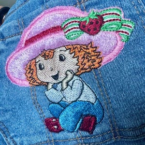 K&ouml;nnte beinhalten: Gestickter Aufn&auml;her mit einer Zeichentrickfigur, die einen rosa Hut mit einer Erdbeere und einer Schleife tr&auml;gt. Die Figur hat orangefarbenes Haar, ein hellblaues Hemd und blaue Hosen. Der Aufn&auml;her ist auf einen blauen Jeansstoff gen&auml;ht.