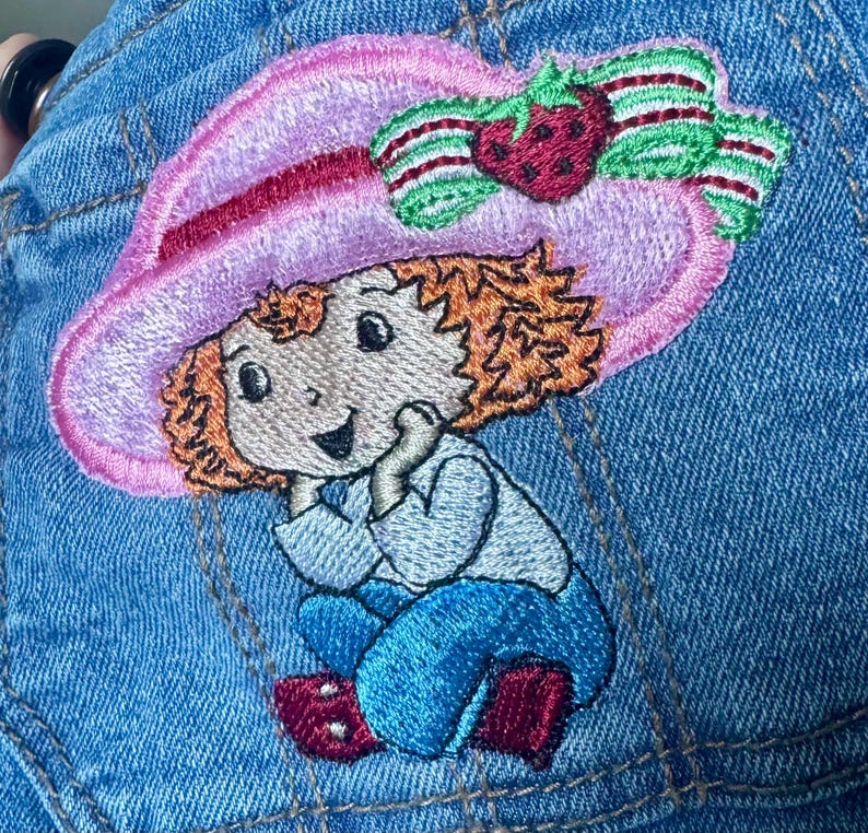 K&ouml;nnte beinhalten: Gestickter Aufn&auml;her mit einer Zeichentrickfigur, die einen rosa Hut mit einer Erdbeere und einer Schleife tr&auml;gt. Die Figur hat orangefarbenes Haar, ein hellblaues Hemd und blaue Hosen. Der Aufn&auml;her ist auf einen blauen Jeansstoff gen&auml;ht.
