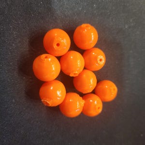 May include: A collection of nine bright orange spherical beads, each featuring a small, centered hole for threading. The beads have a smooth, glossy finish and are arranged in a cluster, showcasing their vibrant, uniform color and round shape.