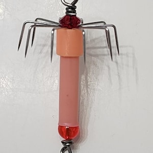 May include: A pink and red squid jig with multiple hooks. The jig has a red bead at the top and bottom and a pink body.