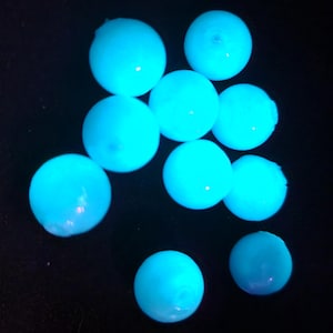 May include: A collection of nine spherical beads that emit a bright, glowing cyan light against a dark background. Each bead is smooth and round, featuring a small central hole, and they are arranged in a loose cluster, showcasing their luminescence.