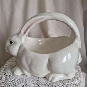 May include: A glossy white ceramic basket shaped like a sitting rabbit. The rabbit has a smooth finish, a black eye, and a handle formed by its long ears that arch over the centre. The inner side of one ear features a soft pink accent.