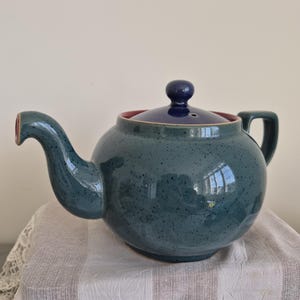 May include: A ceramic teapot with a speckled teal blue glaze, featuring a curved spout, a handle, and a removable lid with a dark blue knob. The interior rim of the teapot is a contrasting reddish-pink colour, and the lid has a small steam vent.