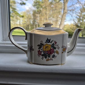 May include: A white porcelain teapot with a gold rim and accents. The teapot features a floral design with yellow, red, and blue flowers. The teapot is sitting on a white windowsill.