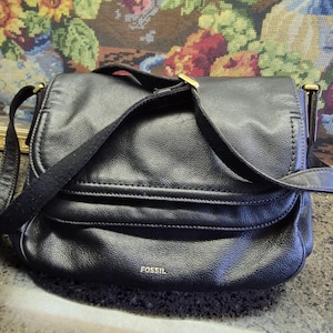 May include: Black leather crossbody bag with a flap closure and adjustable strap. The bag features a gold-tone buckle and the brand name "FOSSIL" embossed on the front. The bag is set against a floral tapestry background.