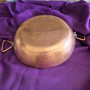 May include: A copper cooking pot with two small handles. The pot has a rounded shape and a shiny surface. The pot is resting on a purple cloth.