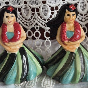 May include: A pair of ceramic salt and pepper shakers shaped like hula dancers. Each figure has black hair with a red flower, a red lei, and a skirt featuring alternating vertical stripes of dark green, light green, and light blue colours.