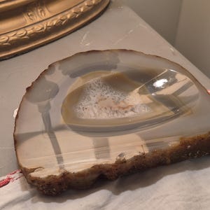 Peut inclure: Une tranche d'agate polie en forme de demi-cercle présentant une surface translucide aux bandes gris clair et blanches, avec une cavité géode centrale contenant de petits cristaux blancs scintillants et un bord extérieur brun naturel.