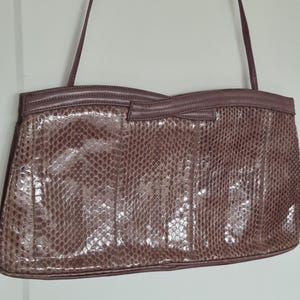 May include: A mauve-coloured shoulder bag featuring a textured snakeskin-like pattern across the front panel. The bag has a structured rectangular shape with a smooth leather trim along the top edge, a small central bow detail, and a thin shoulder strap.