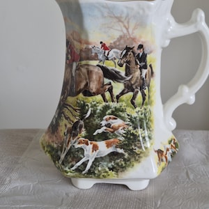 May include: A white ceramic jug featuring a colourful wrap-around scene of a fox hunt. The illustration depicts riders on horseback and several hunting dogs running through a grassy field. The jug has a curved handle and a gold-trimmed rim.