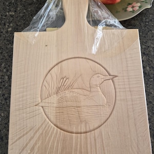 May include: A light-coloured wooden chopping board with a handle. The board features a carved circular design of a loon swimming in water with reeds in the background. The board is wrapped in clear plastic.