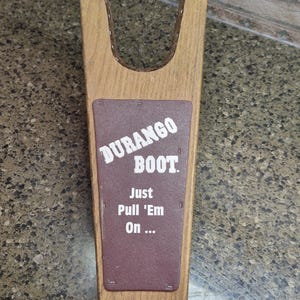 May include: Wooden boot puller with a brown rectangular plate that reads "DURANGO BOOT. Just Pull 'Em On...". The puller is made of light brown wood with a dark brown plate.
