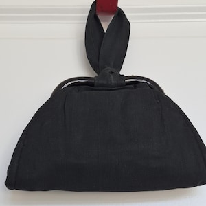 May include: A black fabric handbag featuring a curved dark frame and a single integrated fabric handle loop. The bag has a structured, semi-circular shape with a smooth, matte finish and a clean, minimalist design without any visible external hardware.