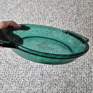 May include: A round, emerald green glass baking dish with integrated side handles. The centre of the dish features embossed text that reads: Anchor Hocking, Fire-King, Oven Ware, Made in U.S.A., 441, 8 INCH, 20 CM, 1.