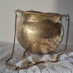May include: A polished brass hanging bowl with a rounded, bulbous body and a wide, scalloped rim. The vessel features three small metal loops attached to the top edge, which connect to a delicate, thin brass chain for suspension purposes.