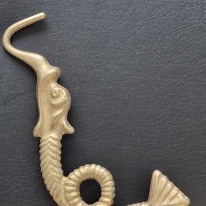 May include: A gold-toned metal hook shaped like a stylised sea creature with a curved neck, a textured body that forms a loop, and a flared tail fin. The object features a smooth, matte finish and a distinct head design with an elongated, hooked top.