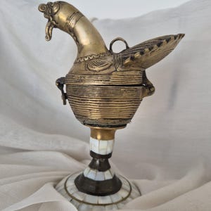 May include: A brass bird-shaped container with etched horizontal line details and a hinged lid. The bird rests on a pedestal base featuring a mosaic pattern of white rectangular mother-of-pearl inlays set against a dark metal frame.