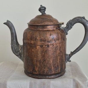 May include: A copper teapot with a dark, weathered patina and a cylindrical body. It features a dark grey metal handle and spout, both decorated with ornate, embossed floral patterns. A matching copper lid with a small, dark, rounded finial sits on top.