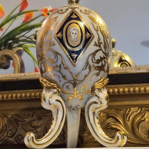 May include: A decorative, egg-shaped porcelain ornament in white with gold detailing and a portrait within a blue diamond. The ornament is supported by a three-legged stand, also with gold trim. The design features intricate gold floral patterns.
