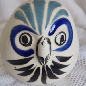 May include: A rounded ceramic owl figurine with an off-white base colour. The owl features hand-painted details including blue arched eyebrows, large black circular eyes, a central black beak, and black feather-like markings below the eyes.