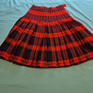 May include: A pleated skirt featuring a vibrant red and black plaid tartan pattern. The garment has a high waistband with a side closure and a flared silhouette that extends from the waist down to the hem, showcasing consistent vertical pleats.